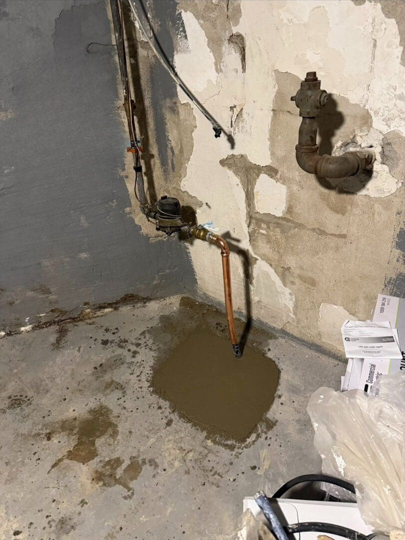 Freshly poured concrete patch on a basement floor near pipes and a damaged wall.