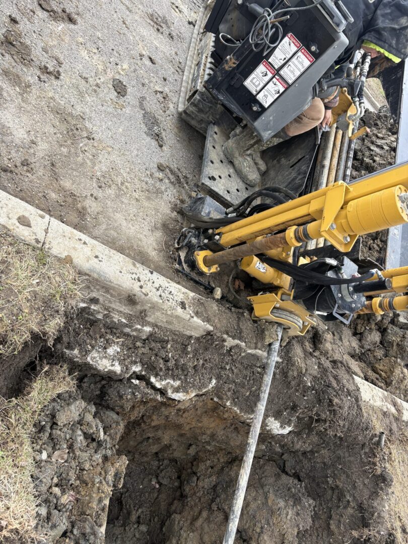Excavator digging a trench near a curb on a construction site.
