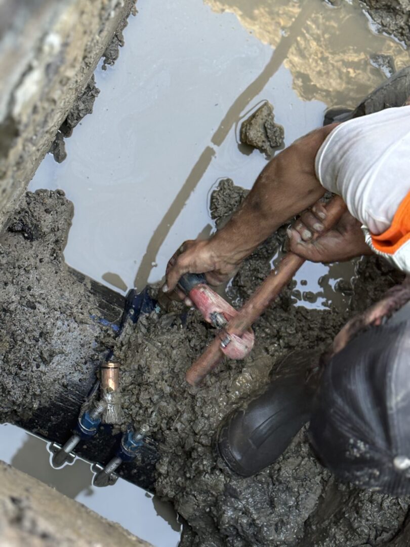 Worker fixing underground water pipe in muddy trench.