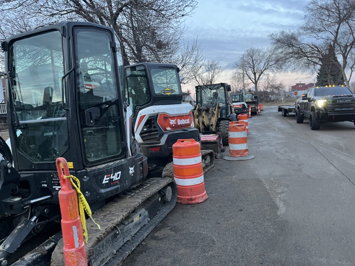 Construction excavators and orange barrels line a road under cloudy skies.
