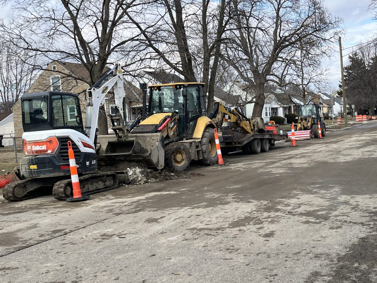 A row of construction vehicles parked along a street lined with bare trees.