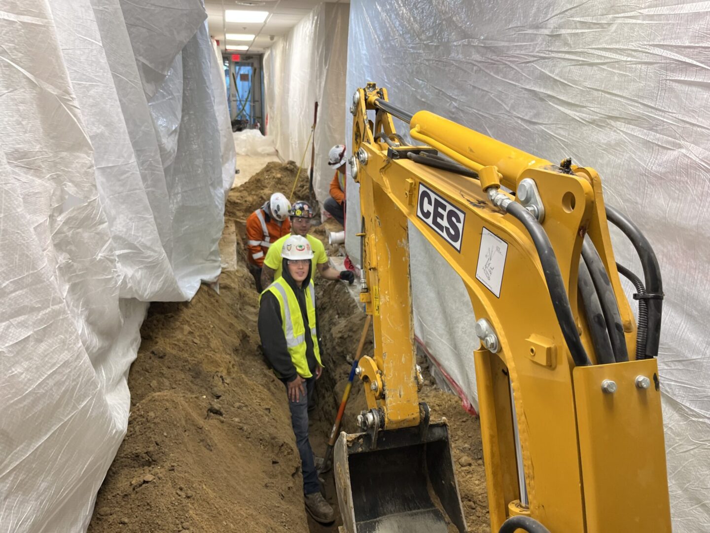 Construction workers operate a JCB excavator in a narrow trench.