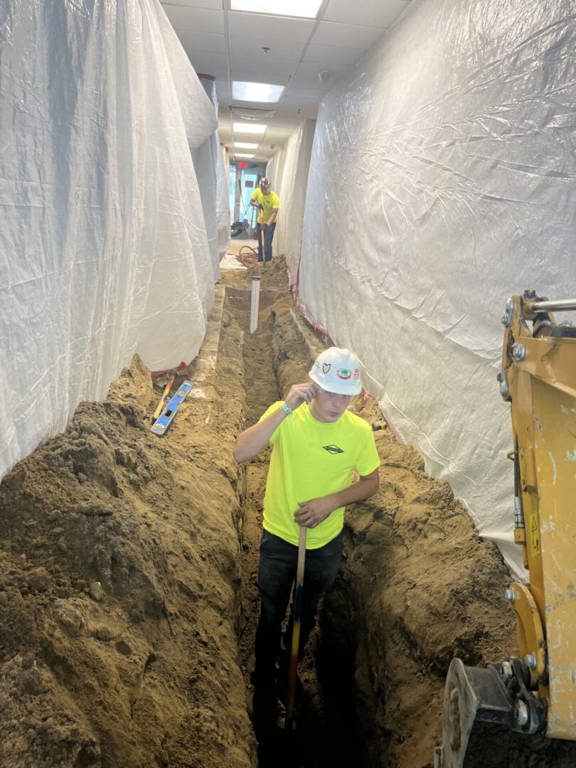Worker in a trench with white walls, wearing a neon shirt and helmet.