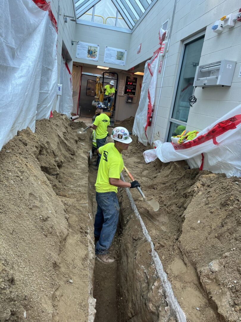 Workers install piping in a deep trench between buildings.