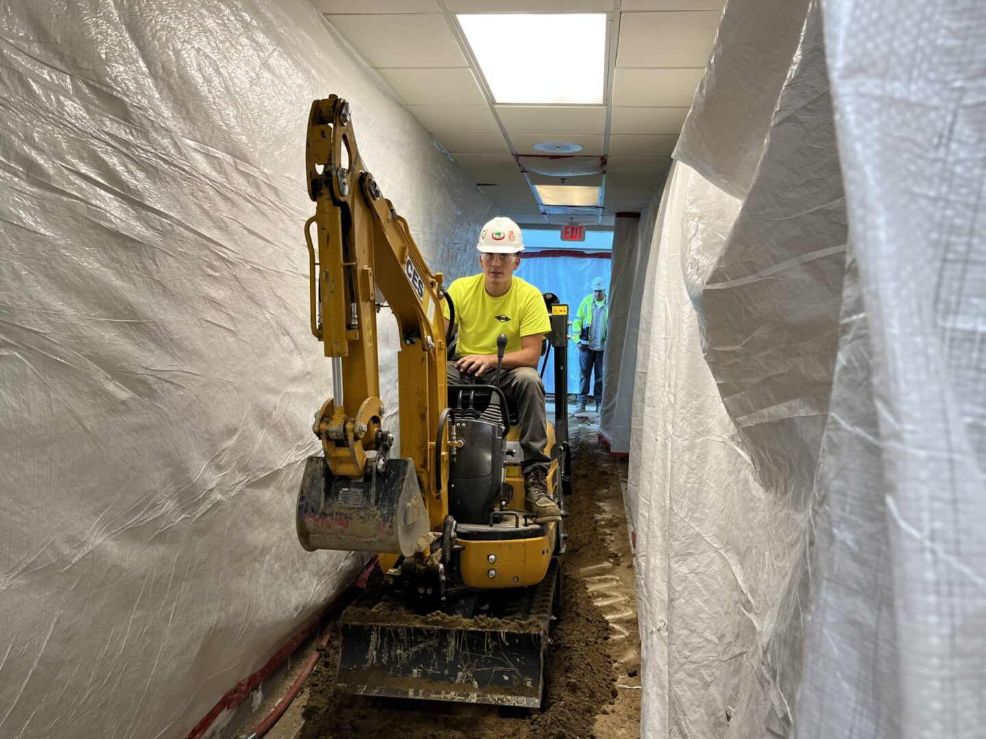 Worker operates mini excavator in narrow indoor construction site.