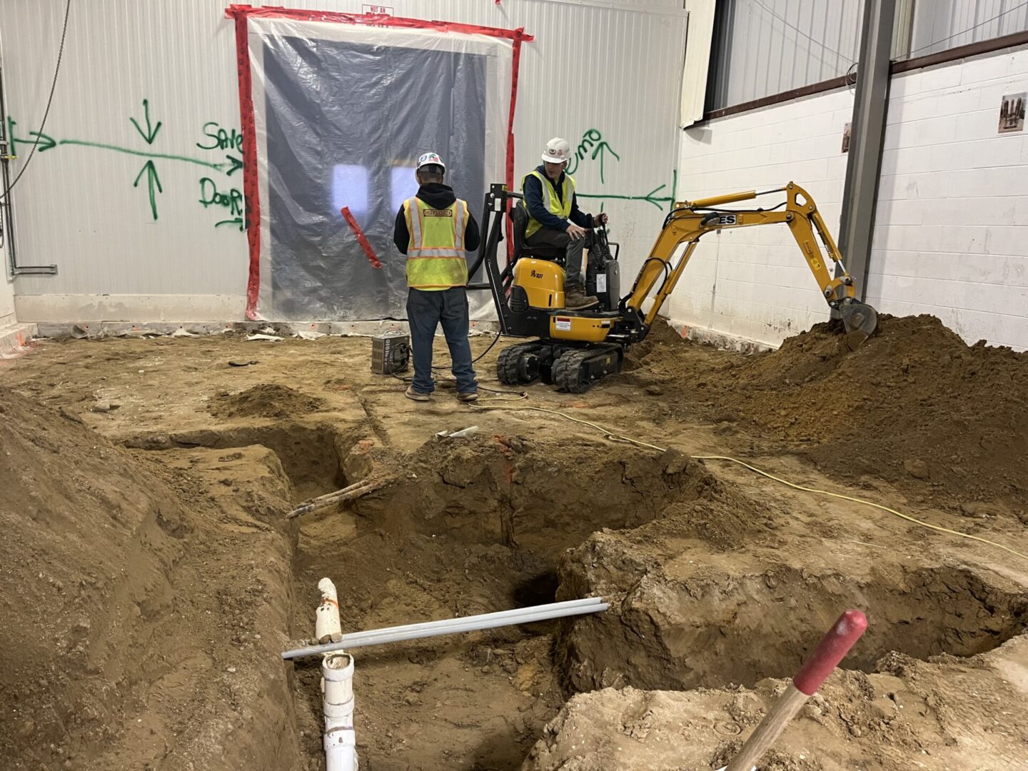Two workers in safety gear operate a mini excavator in an indoor construction site.
