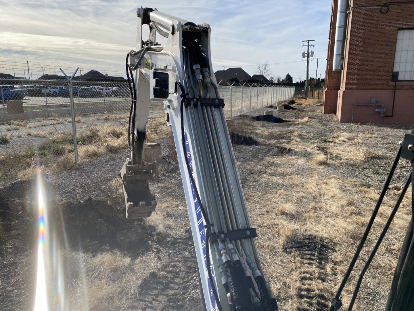 Excavator arm extended over dry, grassy terrain near a brick building.